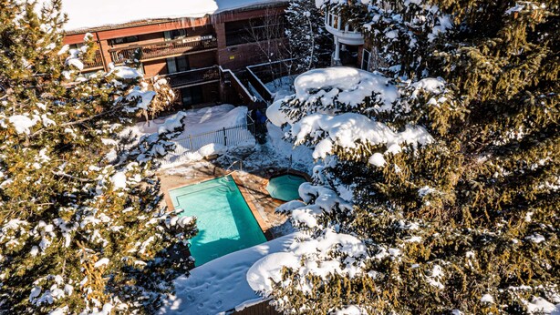 Un área exterior cubierta de nieve con una piscina, rodeada de árboles y un edificio con varios balcones.