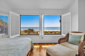 A bedroom with a large window, a bed, a chair, and a view of the beach.