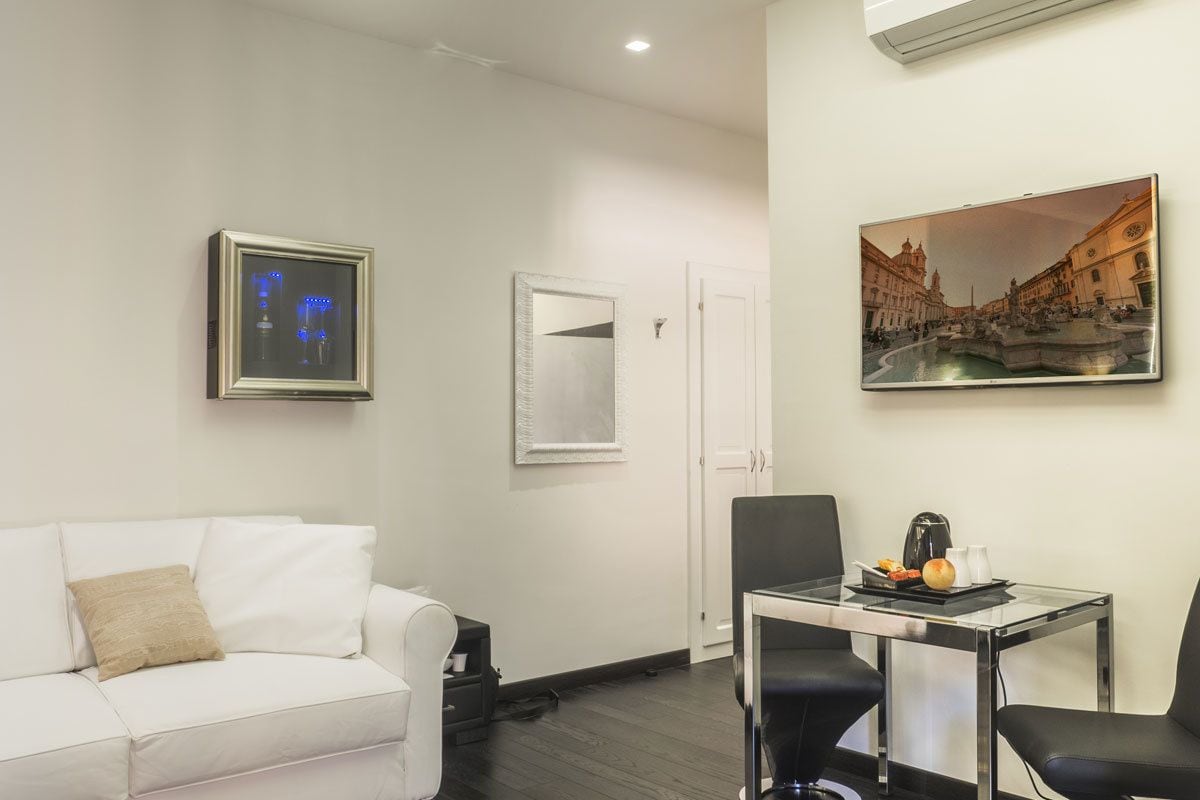 A modern living room with a white sofa, a glass table with two chairs, a framed picture, and a door.