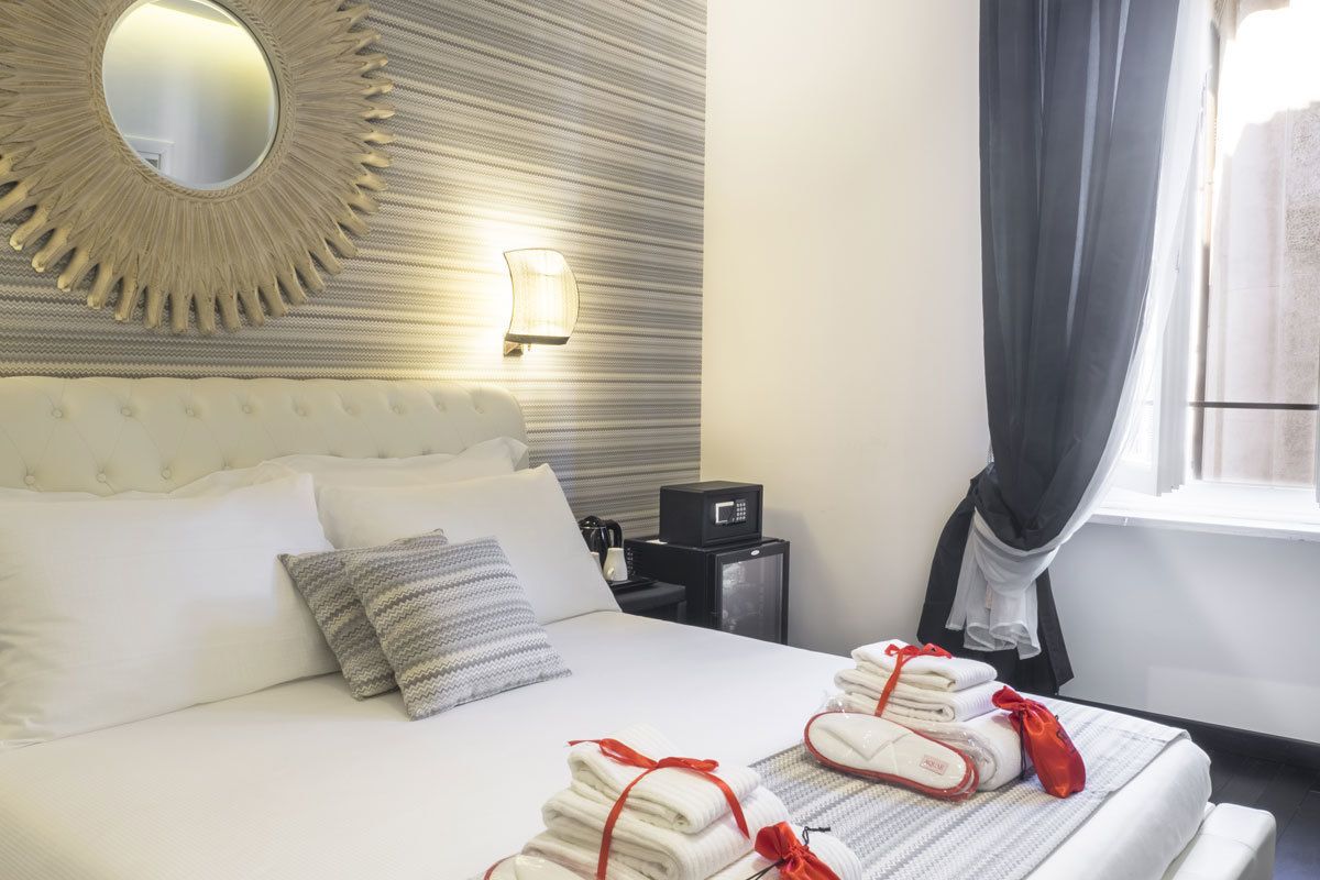 A hotel room with a bed, pillows, a nightstand, a mirror, and a window with curtains.