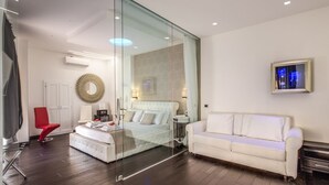 A modern bedroom with a glass partition, a white bed, a red chair, a white sofa, and a framed picture on the wall. - Navona Living Rome (Rome)