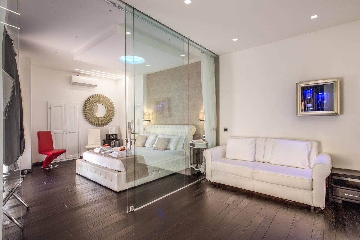 Une chambre moderne avec une cloison en verre, un lit blanc, une chaise rouge, un canapé blanc et un tableau encadré au mur.