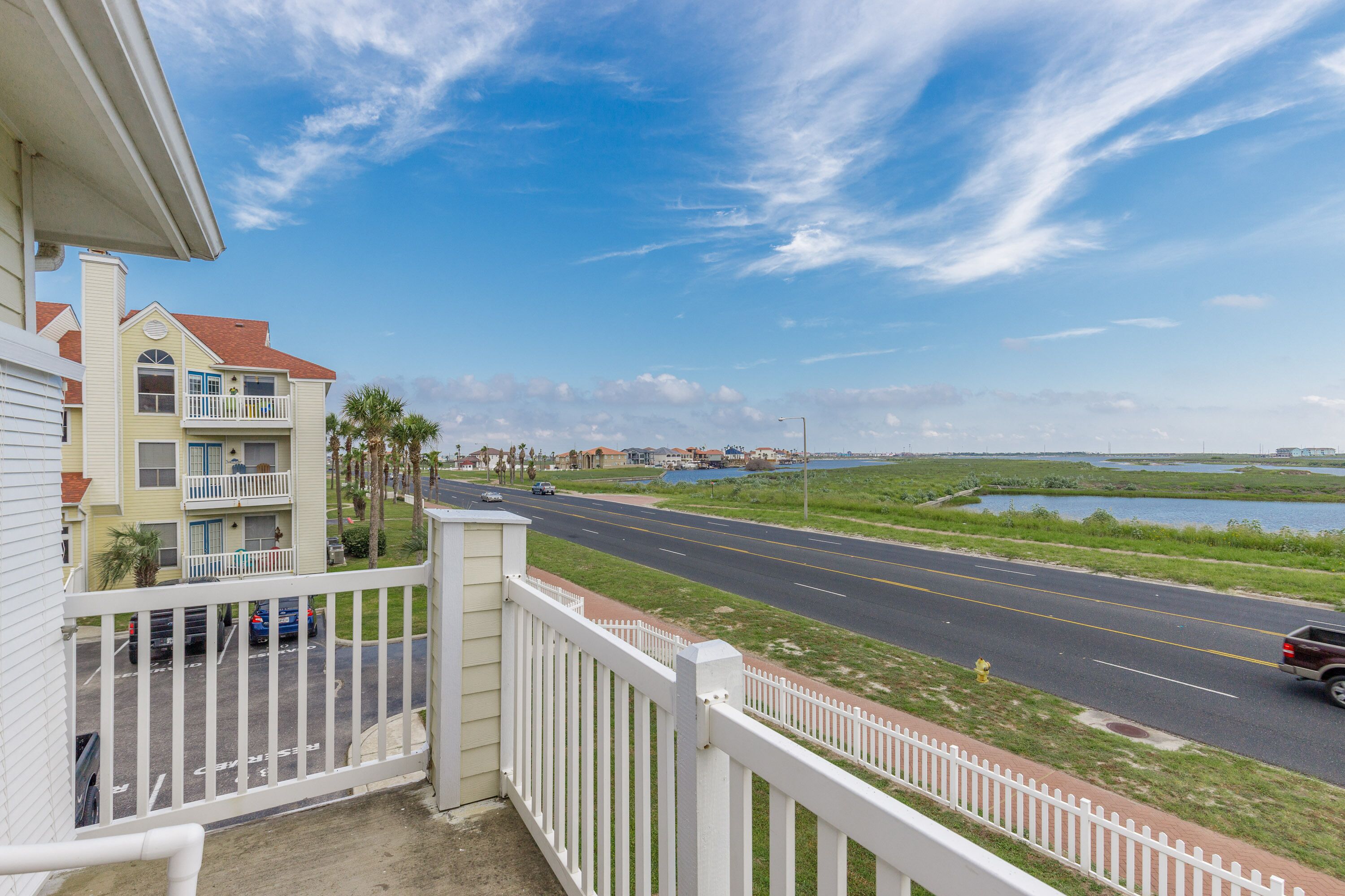 A balcony with a white railing overlooking a road with vehicles, a body of water, and distant buildings.