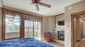A bedroom with a large bed, a fireplace, a television, and a view of trees through the windows.