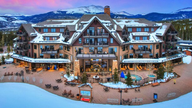 A large hotel with multiple balconies, a central courtyard with outdoor seating, and a mountain backdrop.