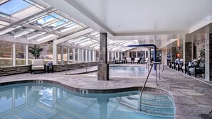 An indoor swimming pool with clear glass ceilings, stone flooring, and seating areas with chairs and tables.