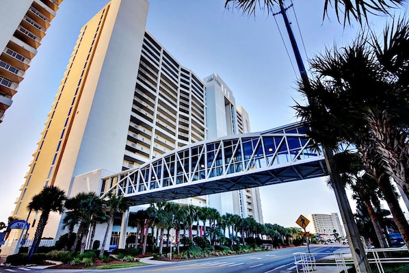 A modern hotel with a prominent glass bridge structure, high-rise buildings, and palm trees in the foreground.