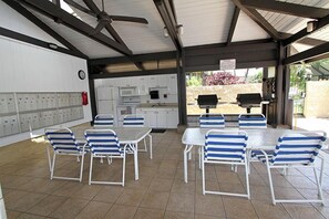 Un espace extérieur abrité, aménagé pour les repas, avec des tables blanches et des chaises à rayures bleues, un coin cuisine doté de meubles blancs et un barbecue.