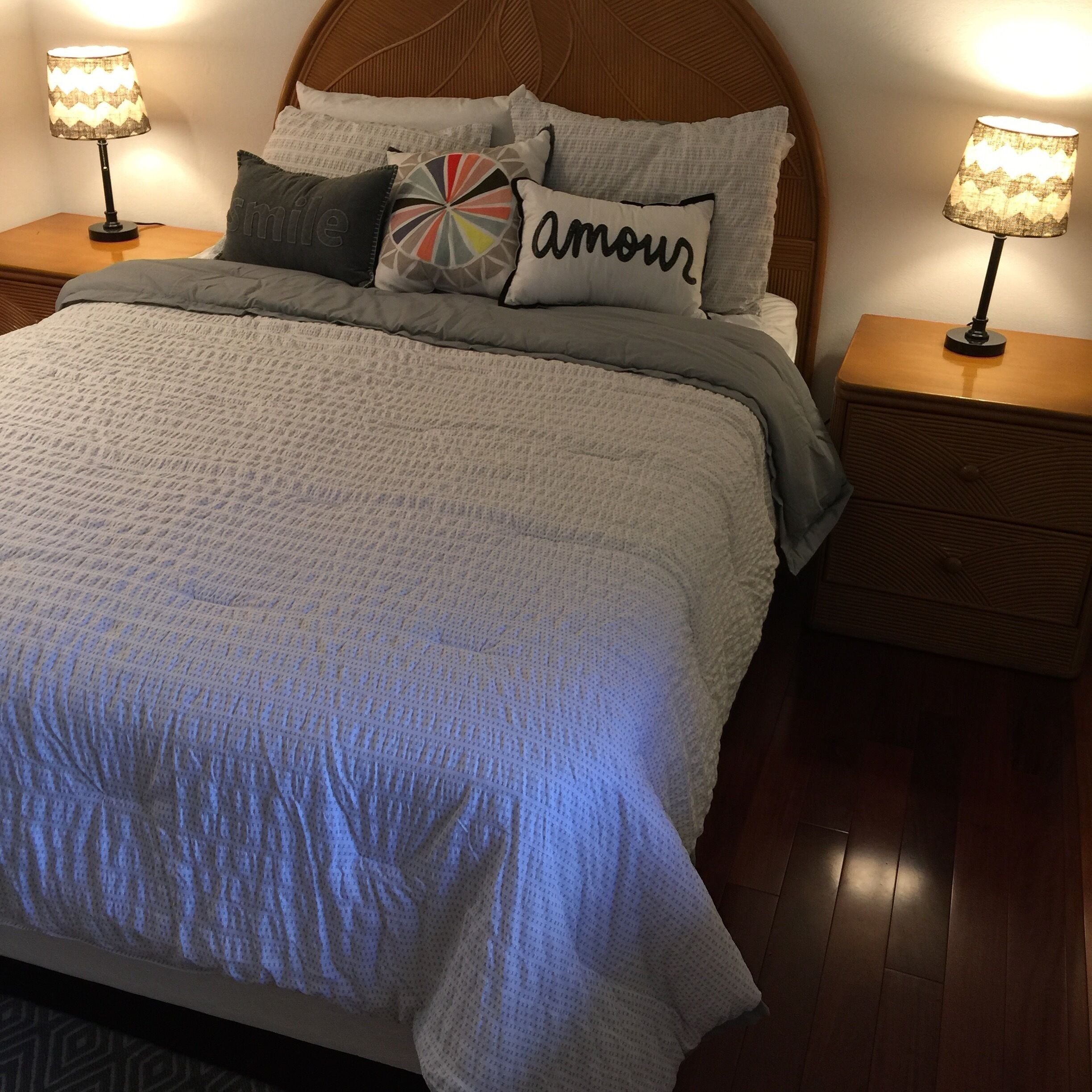 A neatly made bed with a grey quilt, multiple pillows including one with 'amour' written on it, and two bedside lamps on wooden nightstands.