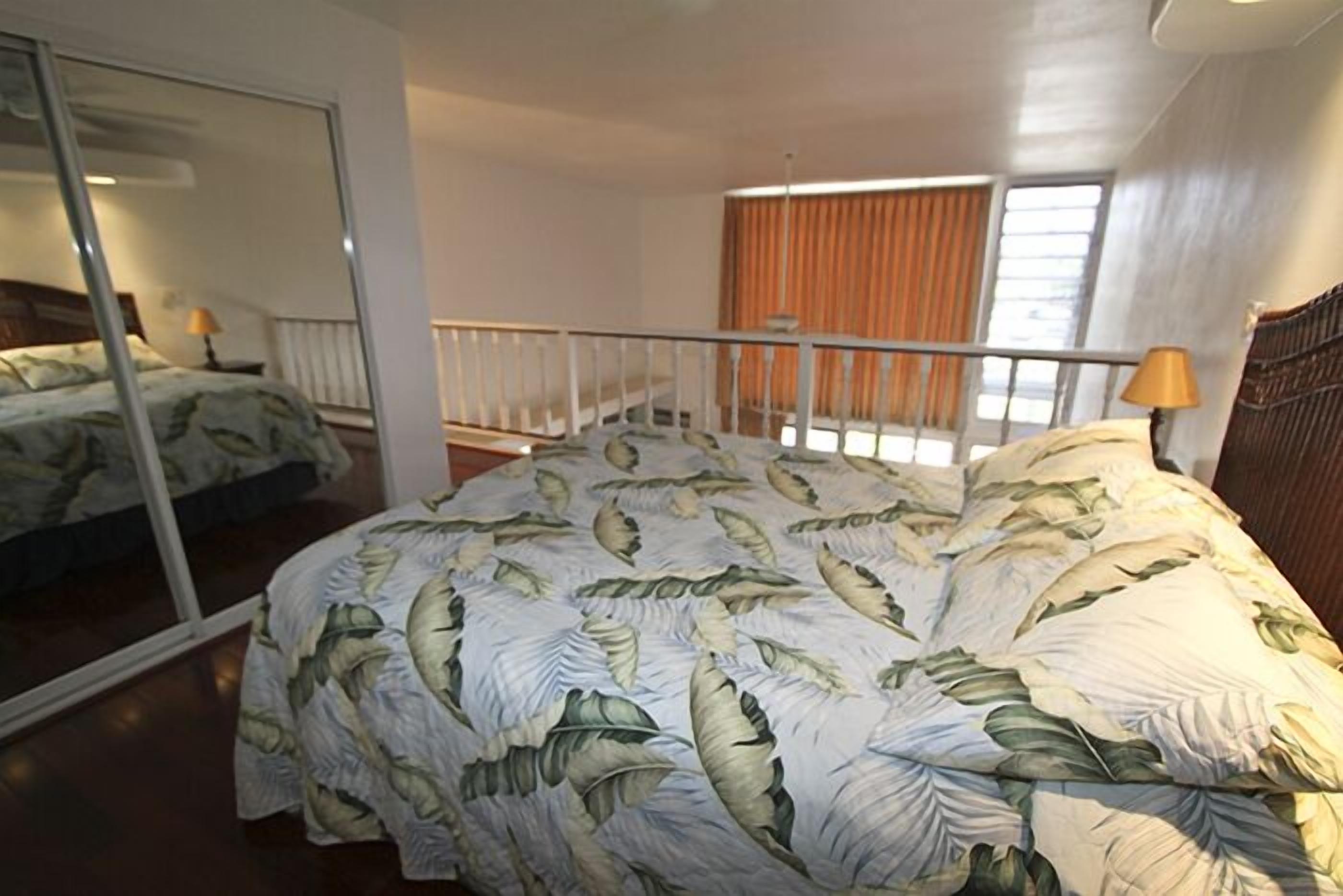 A bedroom with a bed covered in a leaf-patterned duvet, a large mirror, a window with curtains, and a balcony railing.