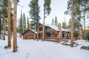Une cabane en rondins avec une cheminée en pierre, entourée de grands pins et d’un sol recouvert de neige.