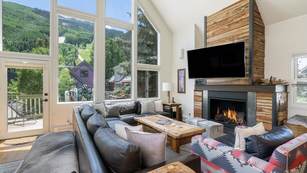 A spacious living room with a large fireplace, comfortable seating, and large windows offering a view of a forested area.