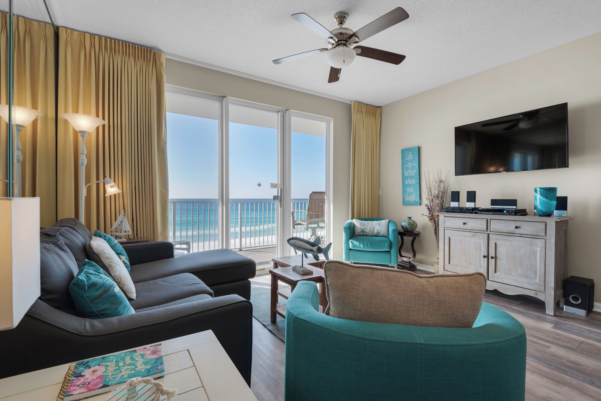 A living room with a sofa, armchair, and a television. There is a ceiling fan, a window with curtains, and a view of the ocean.