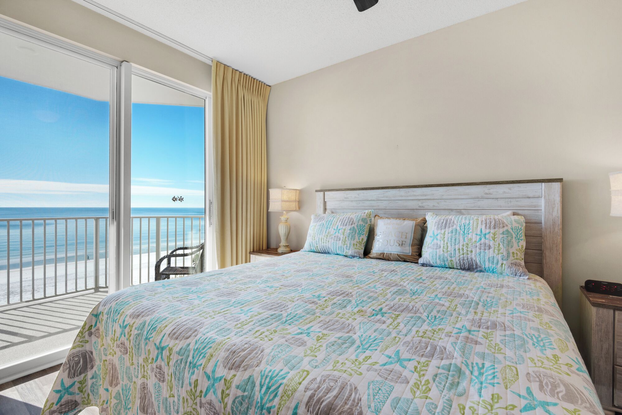 A bedroom with a large bed, a nightstand, a lamp, and a view of the beach.