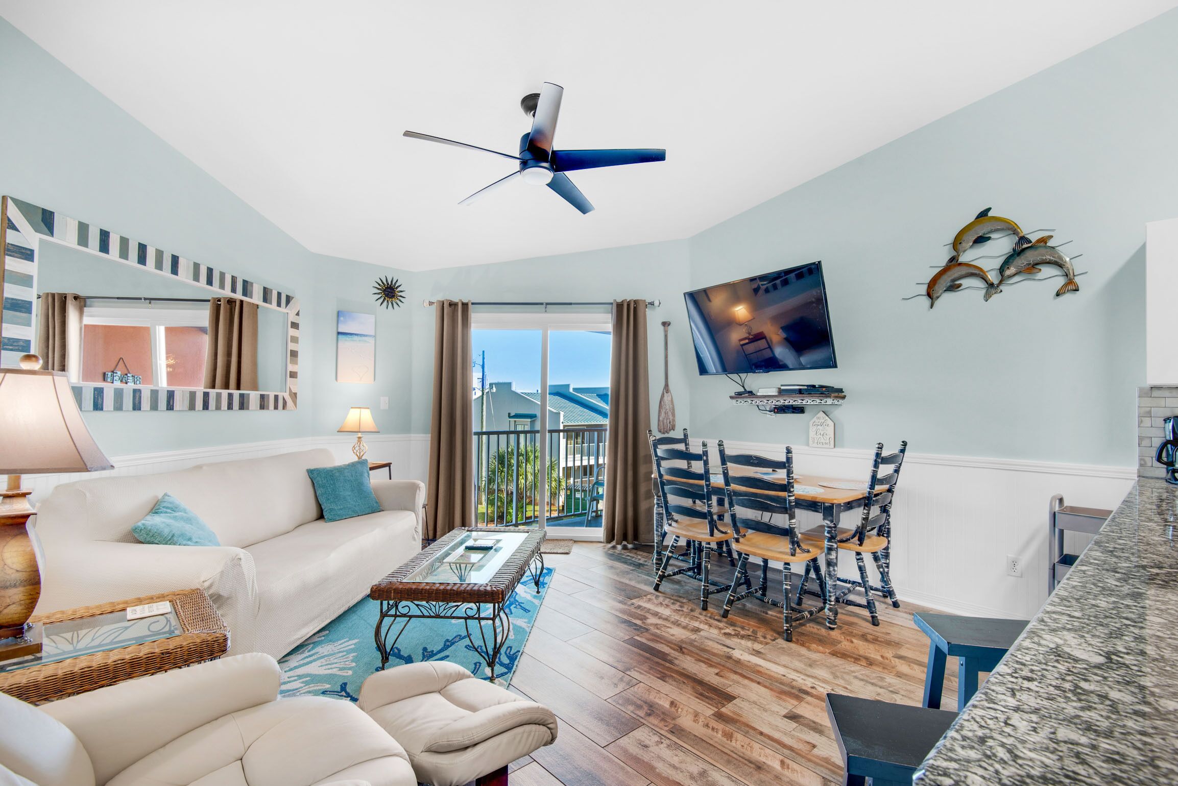 A spacious living room with a sofa, coffee table, and dining area. A ceiling fan and TV are mounted on the wall.