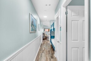 Een hal met houten vloer, lichtblauwe wanden en witte sierlijsten. Aan de rechterkant bevindt zich een deur en in de achtergrond staat een bed met blauw beddengoed.