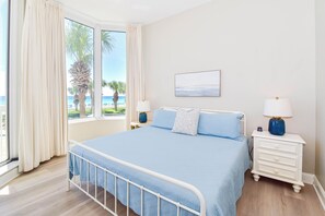 Een slaapkamer met een bed, twee nachtkastjes, een lamp, een schilderij en uitzicht op het strand.