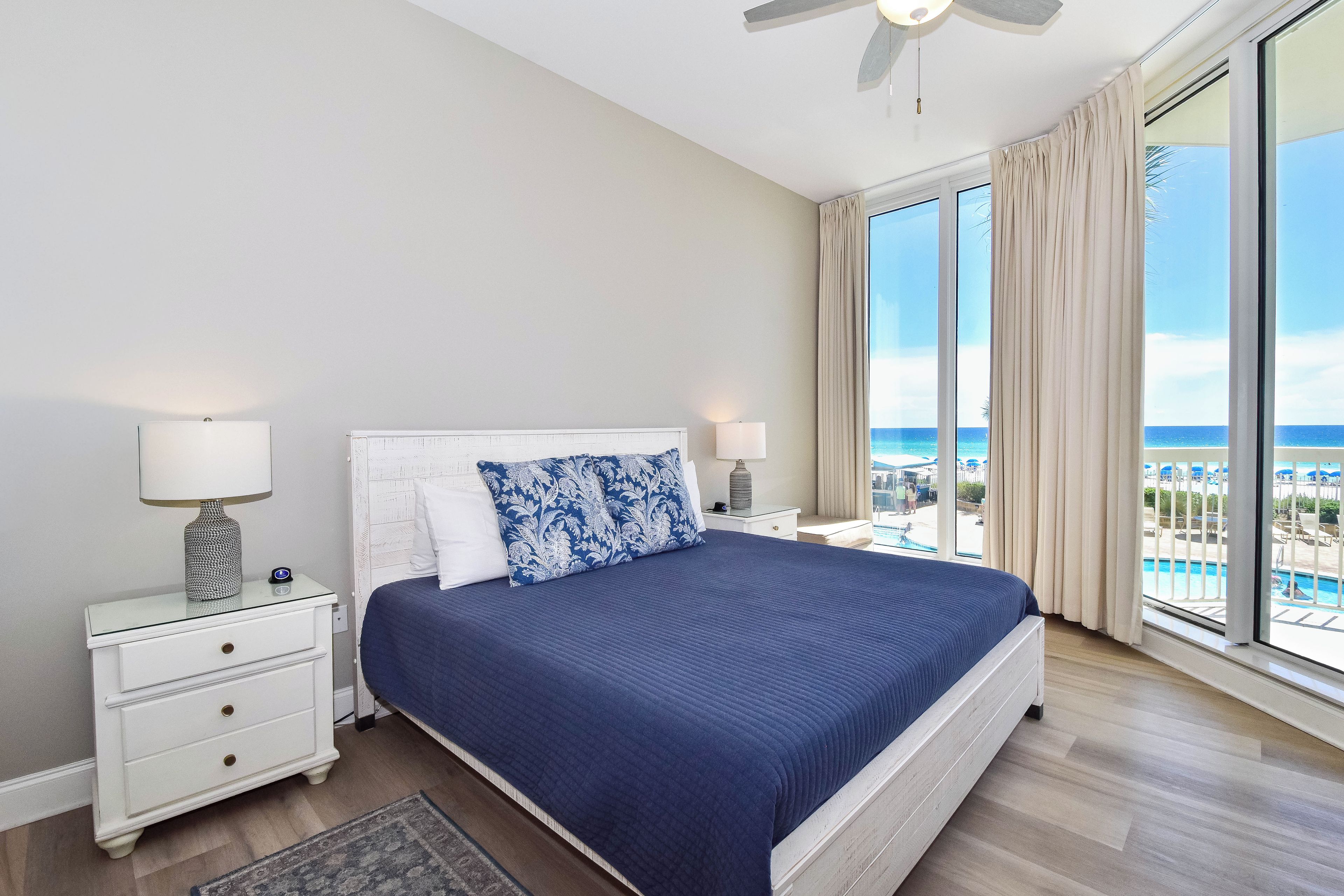 A hotel room with a large bed, two bedside tables, a ceiling fan, and a view of a pool and beach.
