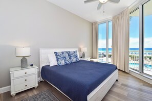 A hotel room with a large bed, two bedside tables, a ceiling fan, and a view of a pool and beach.