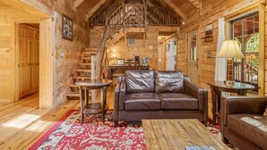 A cozy log cabin interior with a leather sofa, a wooden dining table, and a staircase leading to an upper level.