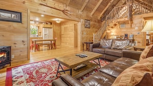 A cozy log cabin interior with a fireplace, leather sofa, and a staircase leading to an upper level.