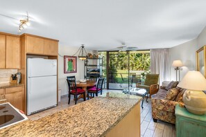 Een keuken met granieten aanrechtbladen, houten keukenkastjes en een inbouwkoelkast. Er is een eethoek met een tafel en stoelen, plus een bank met een lamp. De kamer heeft een grote schuifpui die leidt naar een balkon met uitzicht op groen.