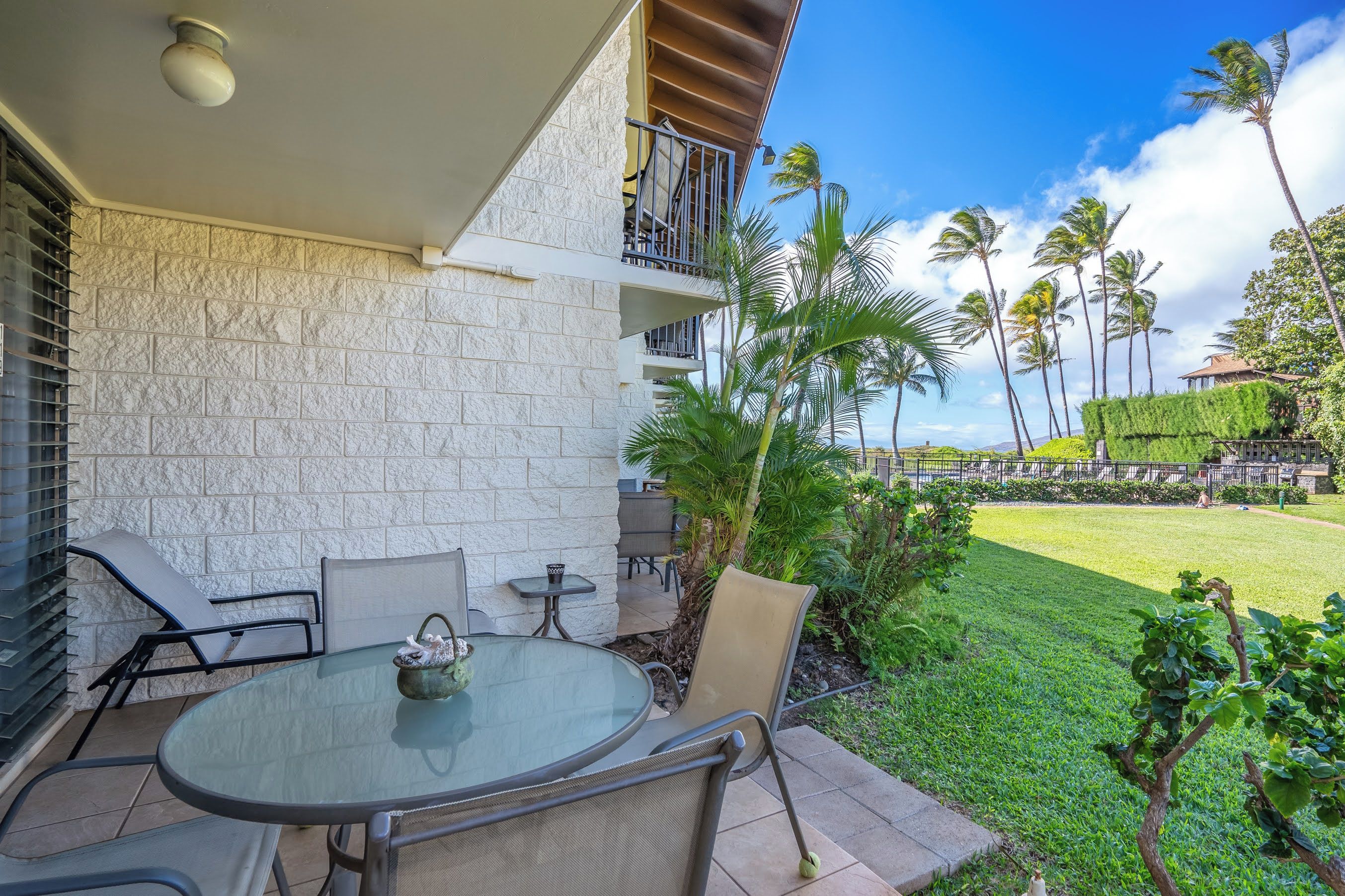 A patio with a round glass table and chairs, a small table with a teapot, and a view of a well-maintained lawn and palm trees.