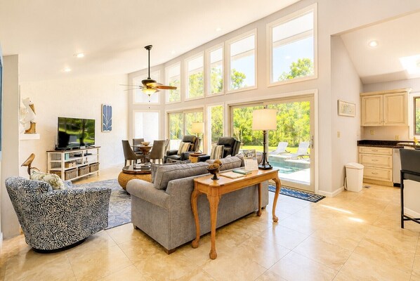 A spacious living room with a large window, a ceiling fan, a television, and a dining area. - 4 Merganser Court -Dog-Friendly, Private Pool, & Quick Walk to Beach/Marina (Hilton Head Island)
