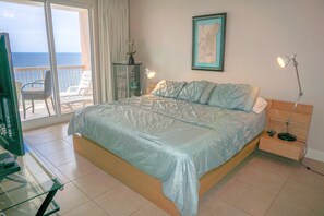 A bedroom with a bed, a nightstand, a lamp, a framed picture on the wall, and a balcony with chairs. - LARGE 3 BEDROOM!! PLENTY OF ROOM & GREAT VIEWS! (Panama City Beach)