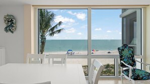 A room with a large window overlooking a beach, a white table, and a chair with a floral pattern.