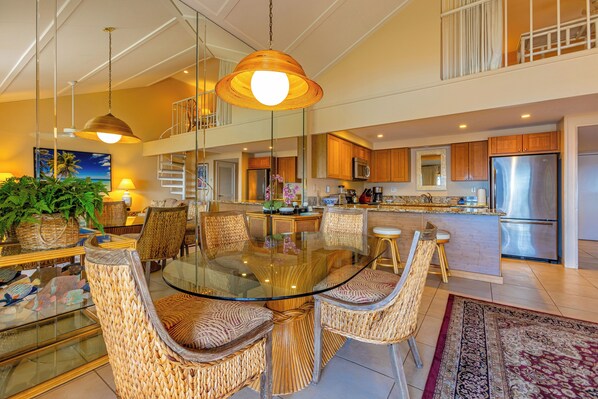 A spacious dining area with a glass-top table surrounded by wicker chairs, a kitchen with stainless steel appliances, and a balcony with a railing. - Ma'alaea Surf #G-7 – Coastal Comfort with Modern Amenities (Kihei)