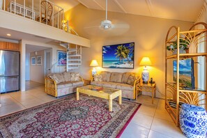 A spacious living room with a high ceiling, a central coffee table, a sofa set, a patterned rug, and a television on a stand. - Ma'alaea Surf #G-7 – Coastal Comfort with Modern Amenities (Kihei)
