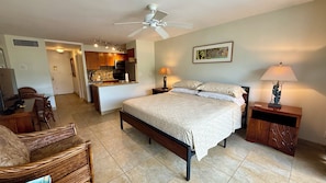 A bedroom with a bed, bedside tables, a lamp, a ceiling fan, a kitchen area, and a dining area.