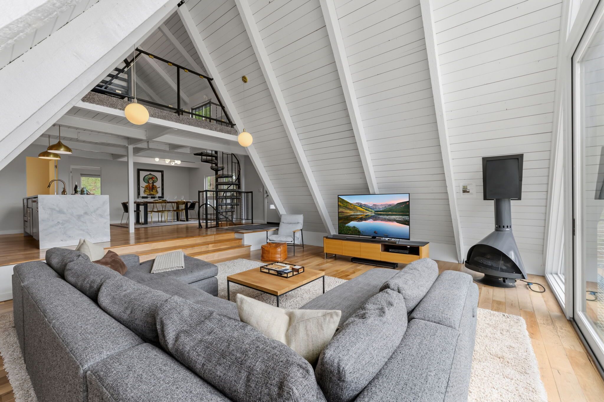 A spacious loft with a high ceiling, a dining area, a living room with a large gray sofa, a fireplace, and a television displaying a scenic landscape.