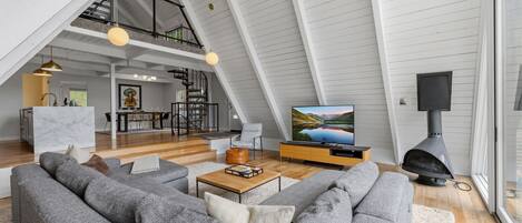 A spacious loft with a high ceiling, a dining area, a living room with a large gray sofa, a fireplace, and a television displaying a scenic landscape.