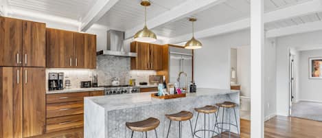 A modern kitchen with wooden cabinets, a marble island, bar stools, and pendant lighting.