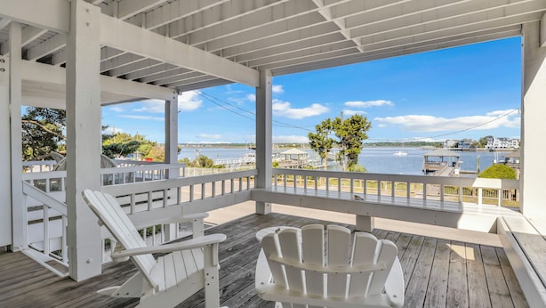 A covered wooden deck with white Adirondack chairs, overlooking a waterfront with buildings and trees. - Conley  A Sound Front Townhome with Boat Slip! (Wrightsville Beach)