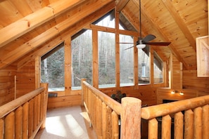 A wooden cabin with a sloped ceiling, large windows, and a ceiling fan.