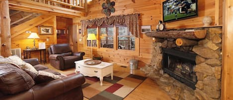 A cozy log cabin interior with a fireplace, leather sofas, a television, and a balcony with a railing.