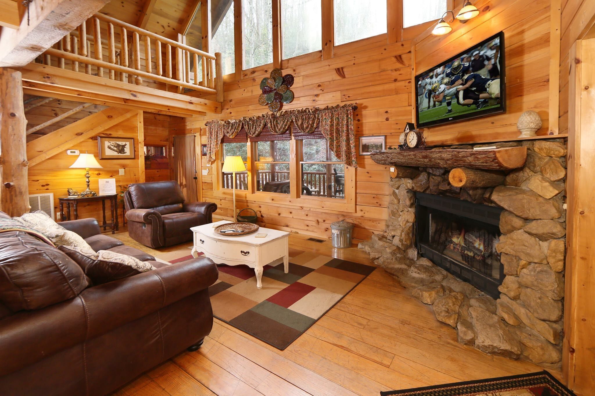 A cozy log cabin interior with a fireplace, leather sofas, a television, and a balcony with a railing.