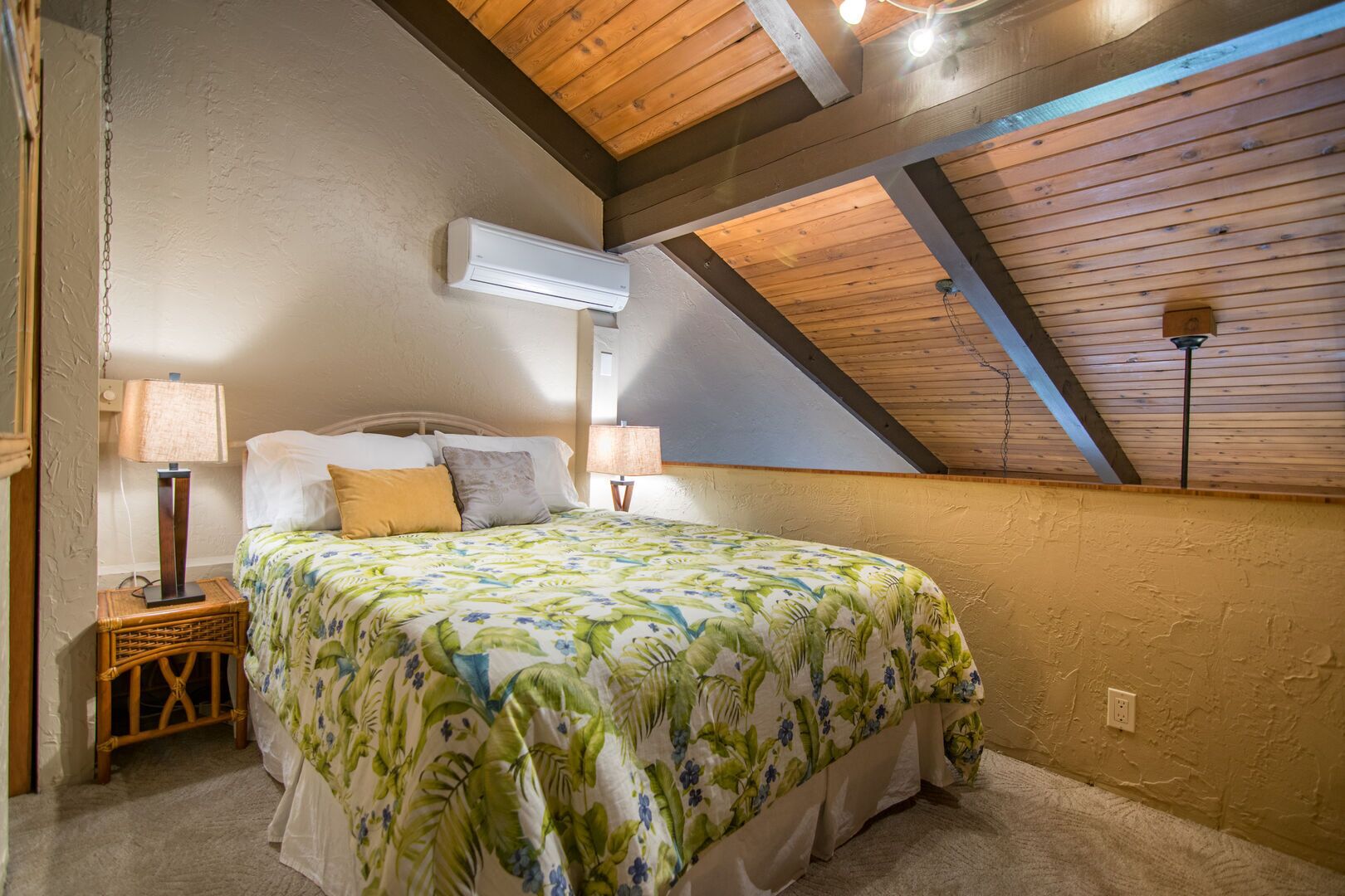 A bedroom with a sloped ceiling, a bed with a floral bedspread, two bedside lamps, and an air conditioning unit.