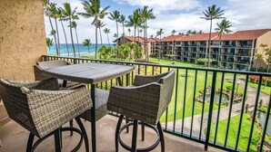 A balcony with wicker furniture overlooking a pool and a hotel building with a red roof. - Remodeled tropical dream with ocean views (Lahaina)