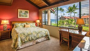 A bedroom with a bed, two bedside tables, a chair, a desk, and a large window with a view of palm trees and a building. - Remodeled tropical dream with ocean views (Lahaina)