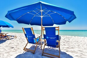 Dos sillas de playa azules con la marca 'La Dolce Vita' sobre una playa arenosa con cielo azul despejado y océano al fondo.