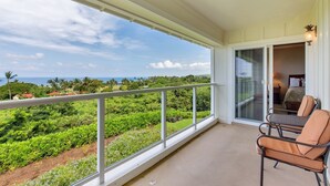 Un balcone con vista su vegetazione rigogliosa e l'oceano. Sono presenti due sedie e un'ampia porta scorrevole in vetro che dà accesso a una camera da letto.