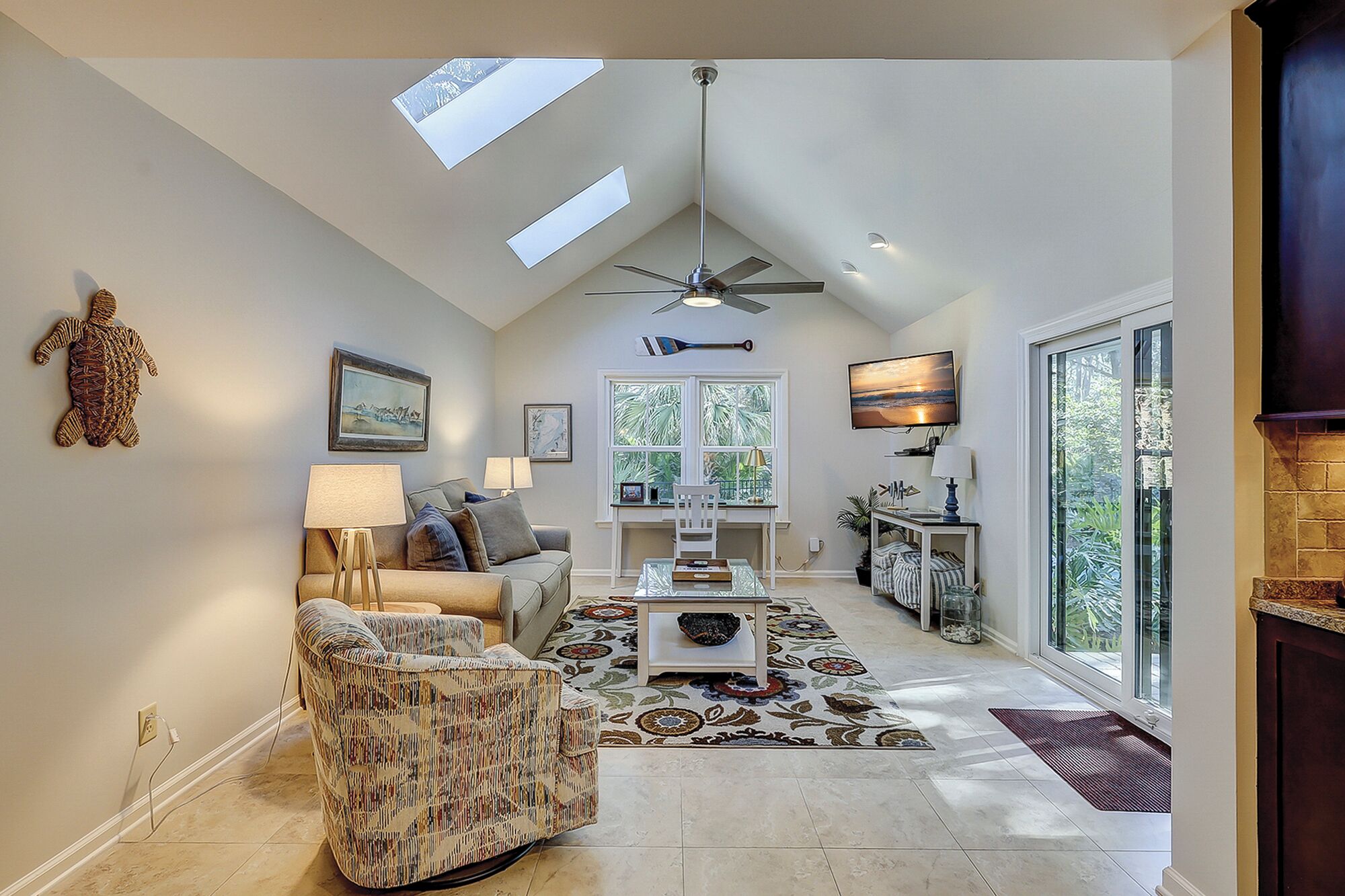 A spacious living room with a high ceiling, skylights, a ceiling fan, a fireplace, and a large window providing a view of the outdoors.