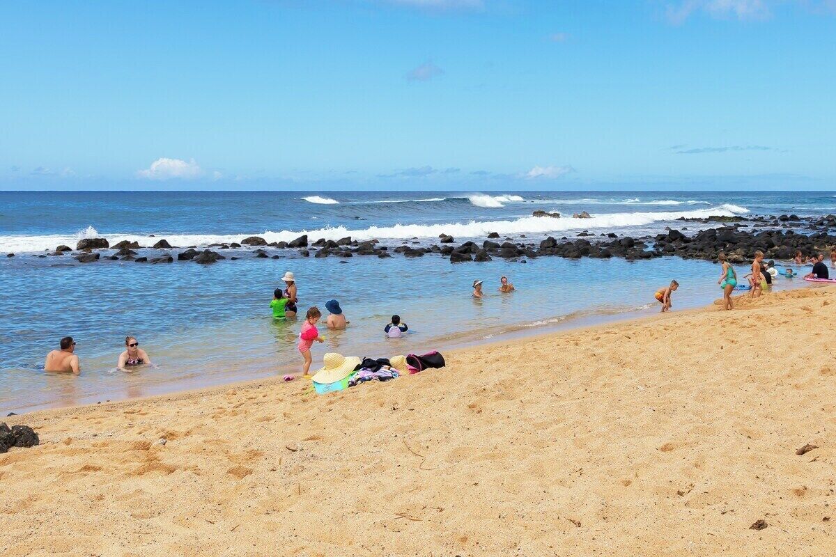 A beach with people swimming and playing in the water, a rocky shoreline, and a sandy beach area with some items on the sand.
