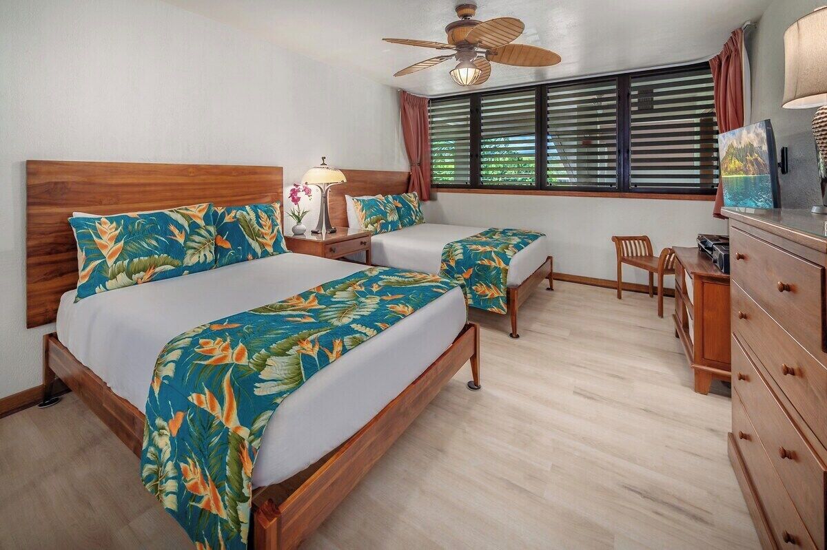 Une chambre d’hôtel avec deux lits, une tête de lit en bois, un ventilateur de plafond, une fenêtre avec des rideaux, un bureau avec une lampe et une commode.
