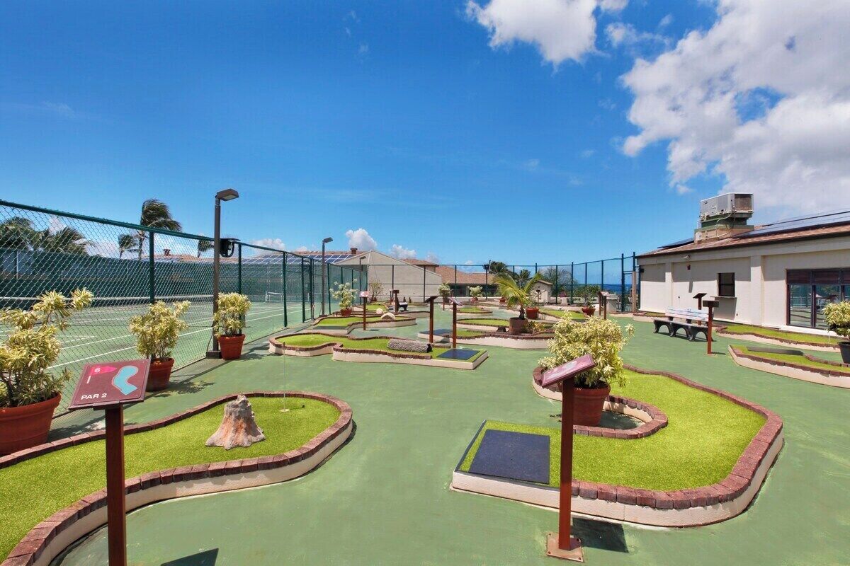 A miniature golf course with a clear path marked by a sign, surrounded by green artificial turf and potted plants.