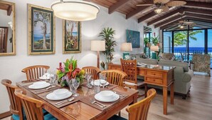 A dining area with a wooden table set for six, surrounded by wooden chairs. There are framed tropical paintings on the wall, a large potted plant, and a view of the ocean through large windows. - Lawai Beach 1401 Oceanfront By Suite Paradise (Koloa)